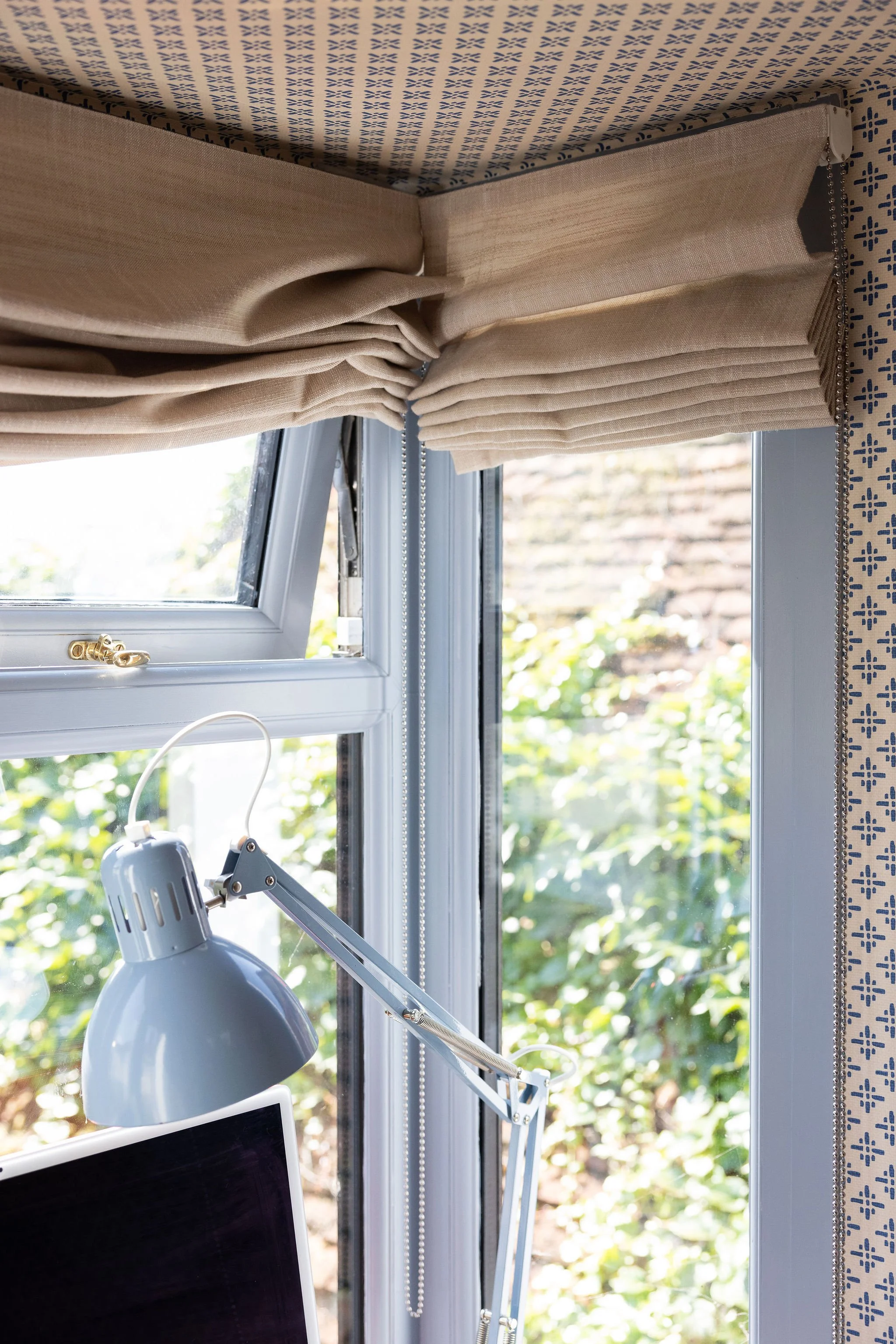 Close-up of a window with beige curtains and a blue patterned valance at the top, with a white desk lamp and part of a computer monitor in the foreground. Outside the window, greenery and a brick wall are visible.