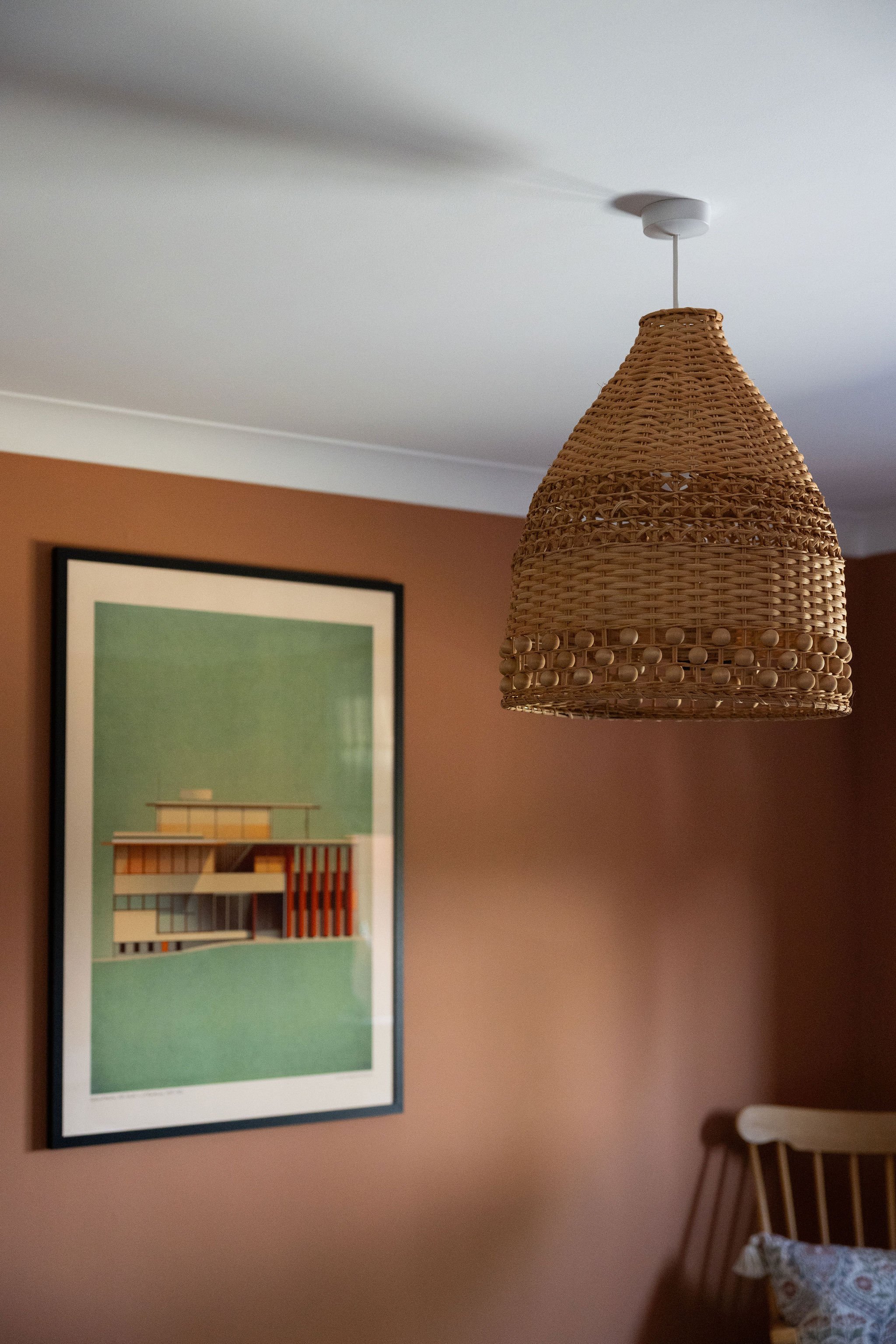 A woven wicker lampshade hanging from the ceiling in a room with orange walls. There is a framed piece of modern artwork on the wall and a partially visible wooden chair with a patterned cushion in the corner.