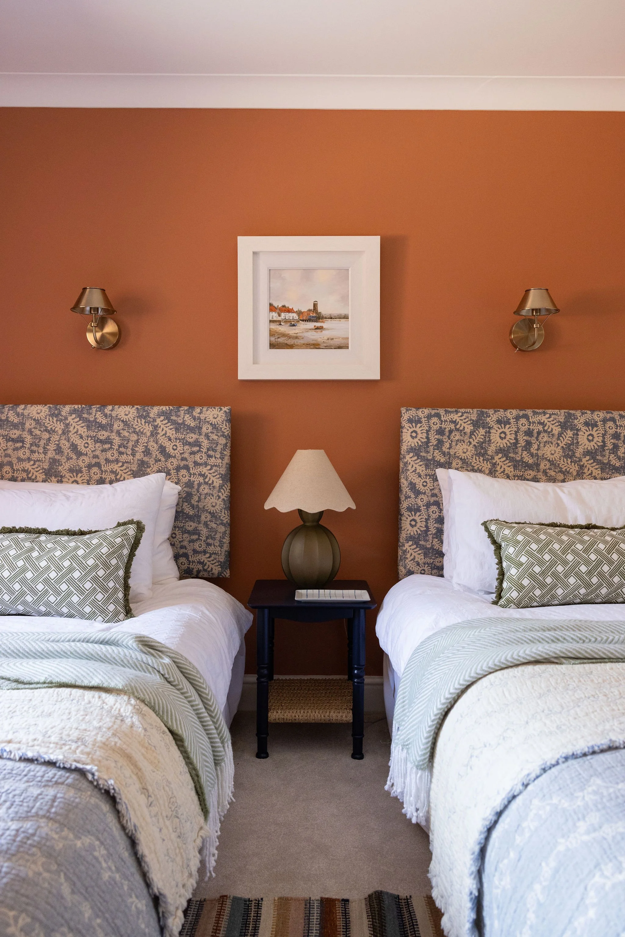 Two beds with patterned headboards and white bedding with green and beige accents, separated by a dark wood nightstand with a lamp, set against an orange wall with framed seaside painting and wall-mounted lamps.