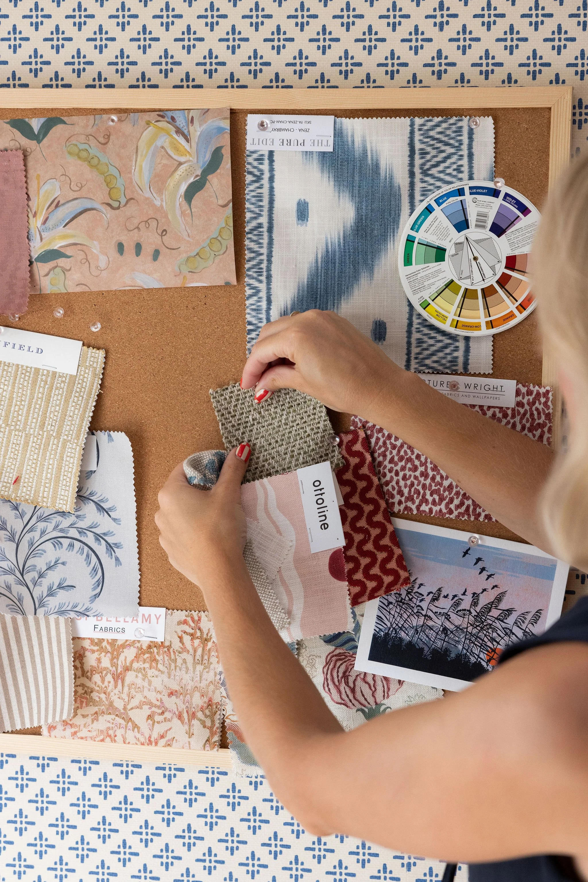 Person selecting fabric samples with various patterns and colors, several fabric swatches and a color wheel on a corkboard, and decorative wallpaper in the background.