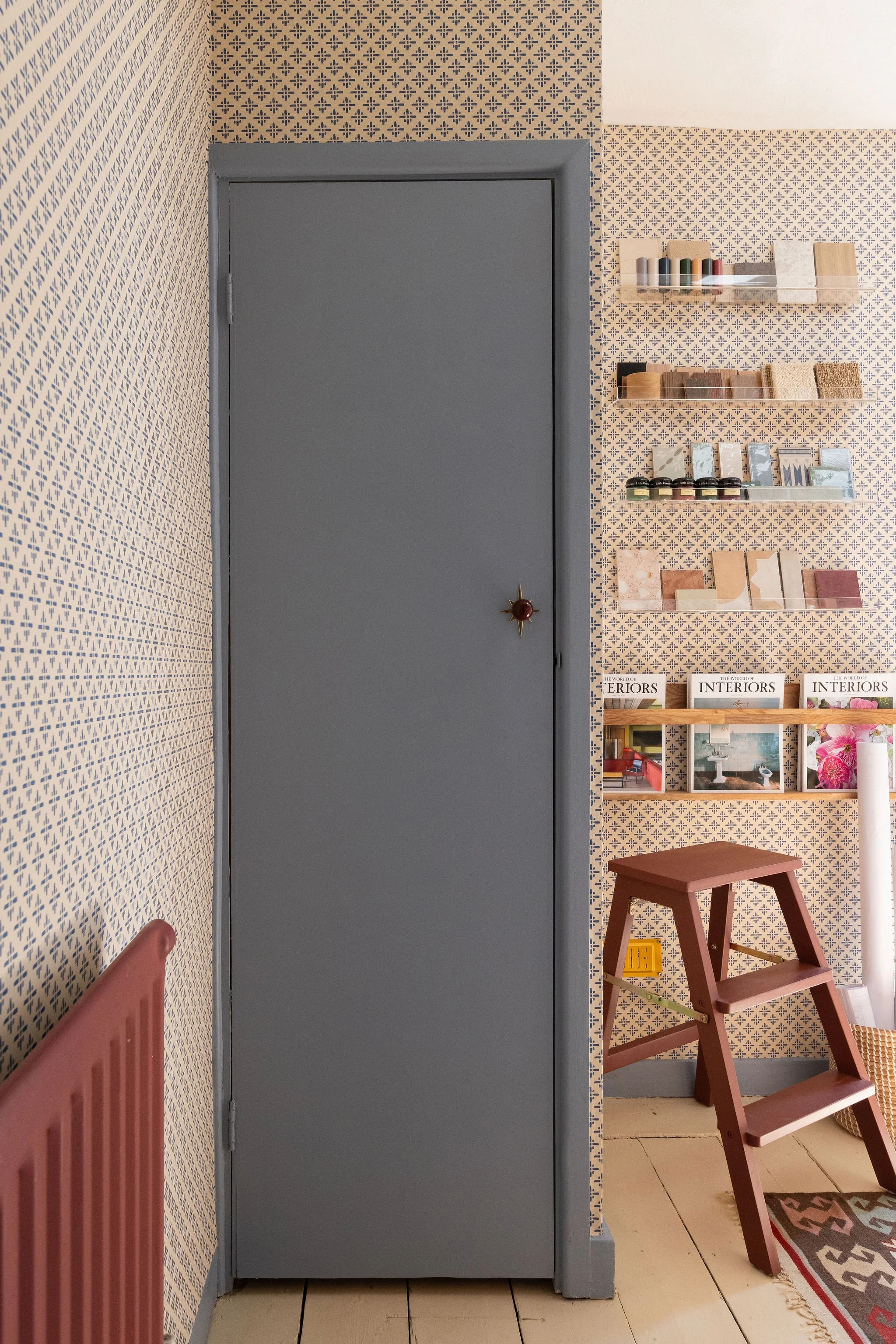 A blue door next to a textured wall with a small open shelf holding colourful spools of thread and fabric samples. A pink wooden step stool sits in front of the wall, and part of a colorful rug is visible on the floor.