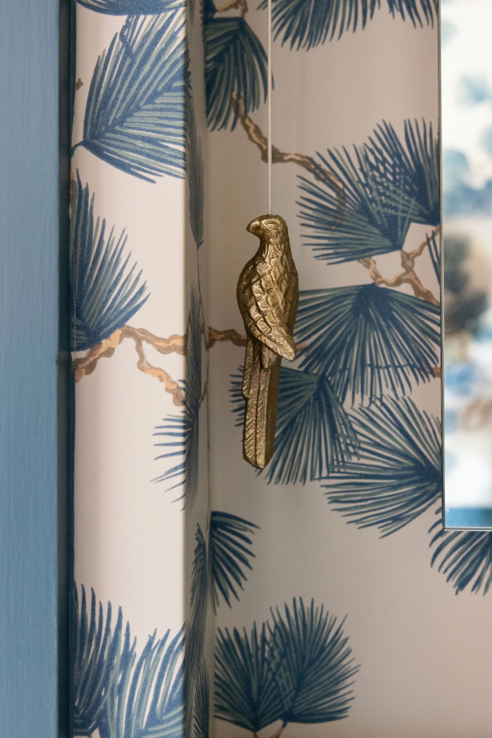 Decorative blue palm leaf pattern on a wall or divider, with a gold-colored bird ornament hanging from a string in front of it.