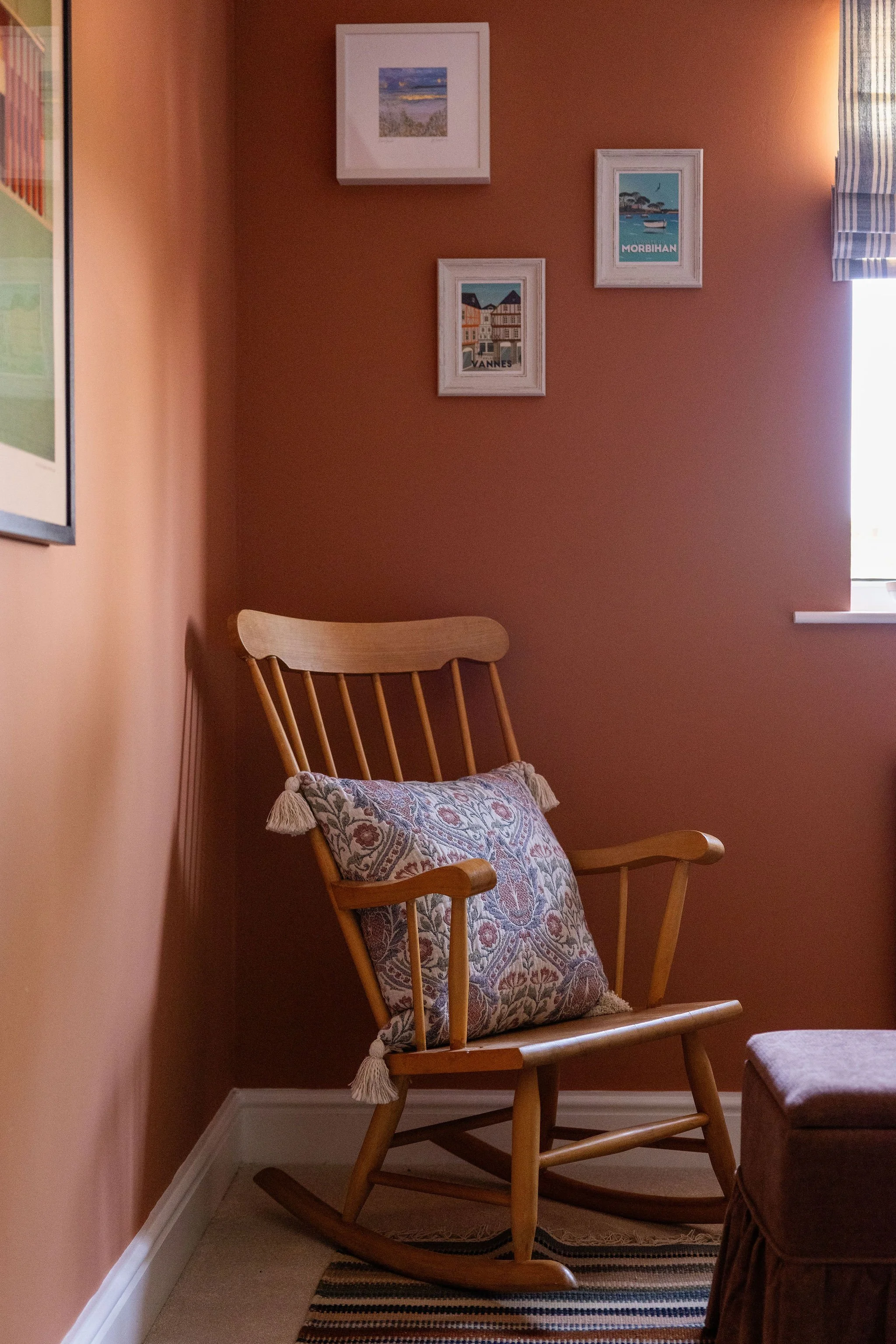 A wooden rocking chair with a decorative cushion, located next to a window with striped blue curtains, in a room with peach-colored walls decorated with framed artwork.