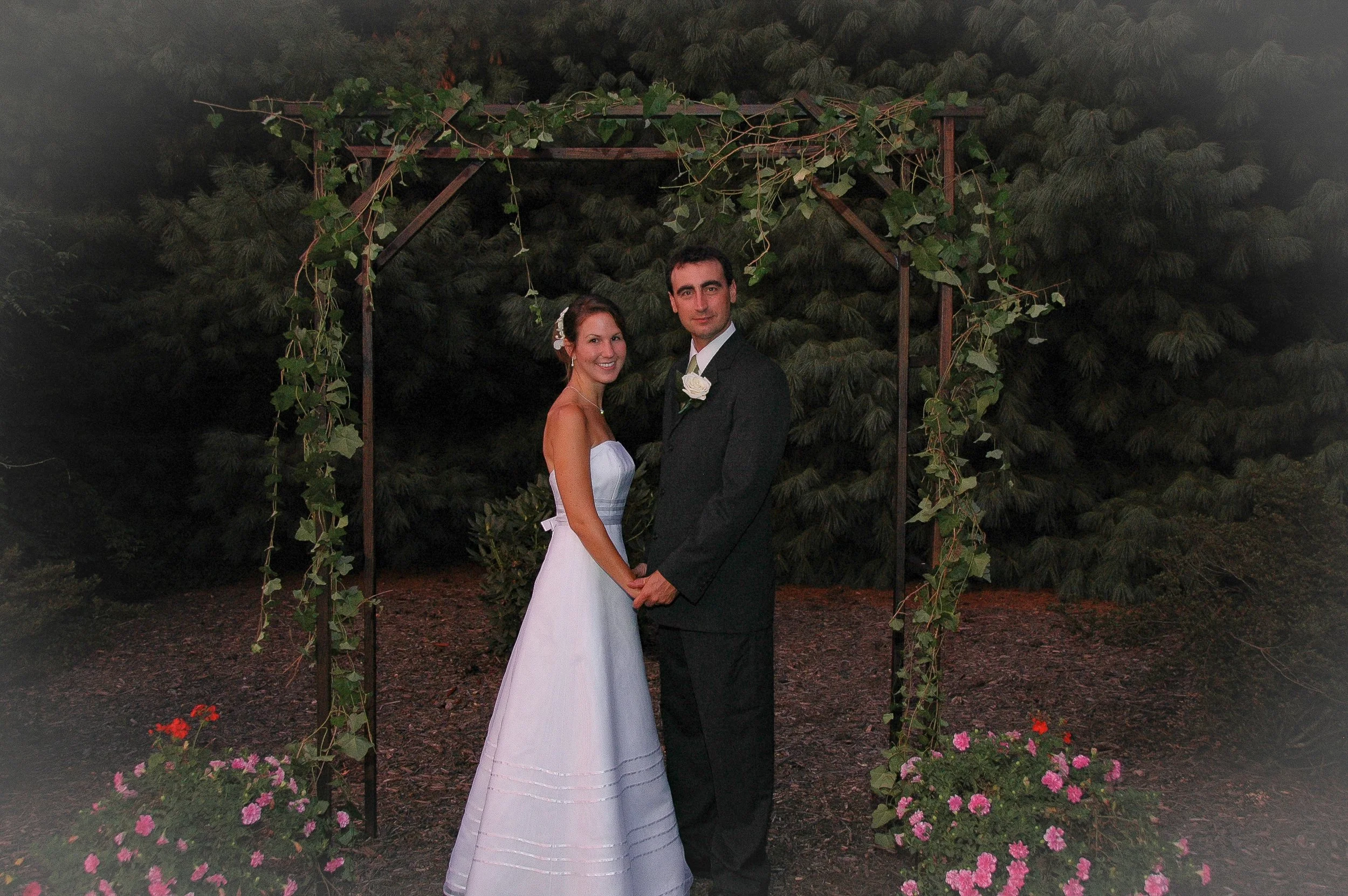 A bride and groom holding hands under a decorative arch with green vines, standing outdoors at night, with trees and flowers in the background.