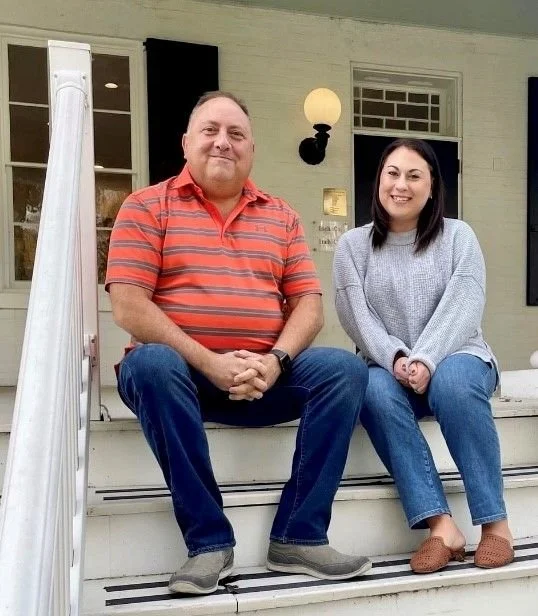 A man and woman sit on the porch steps of a house, smiling at the camera. The man wears a red and gray striped polo shirt, jeans, and gray shoes. The woman wears a gray sweater, jeans, and brown slippers. The house has a light-colored exterior with windows, a door, and a wall-mounted lamp.
