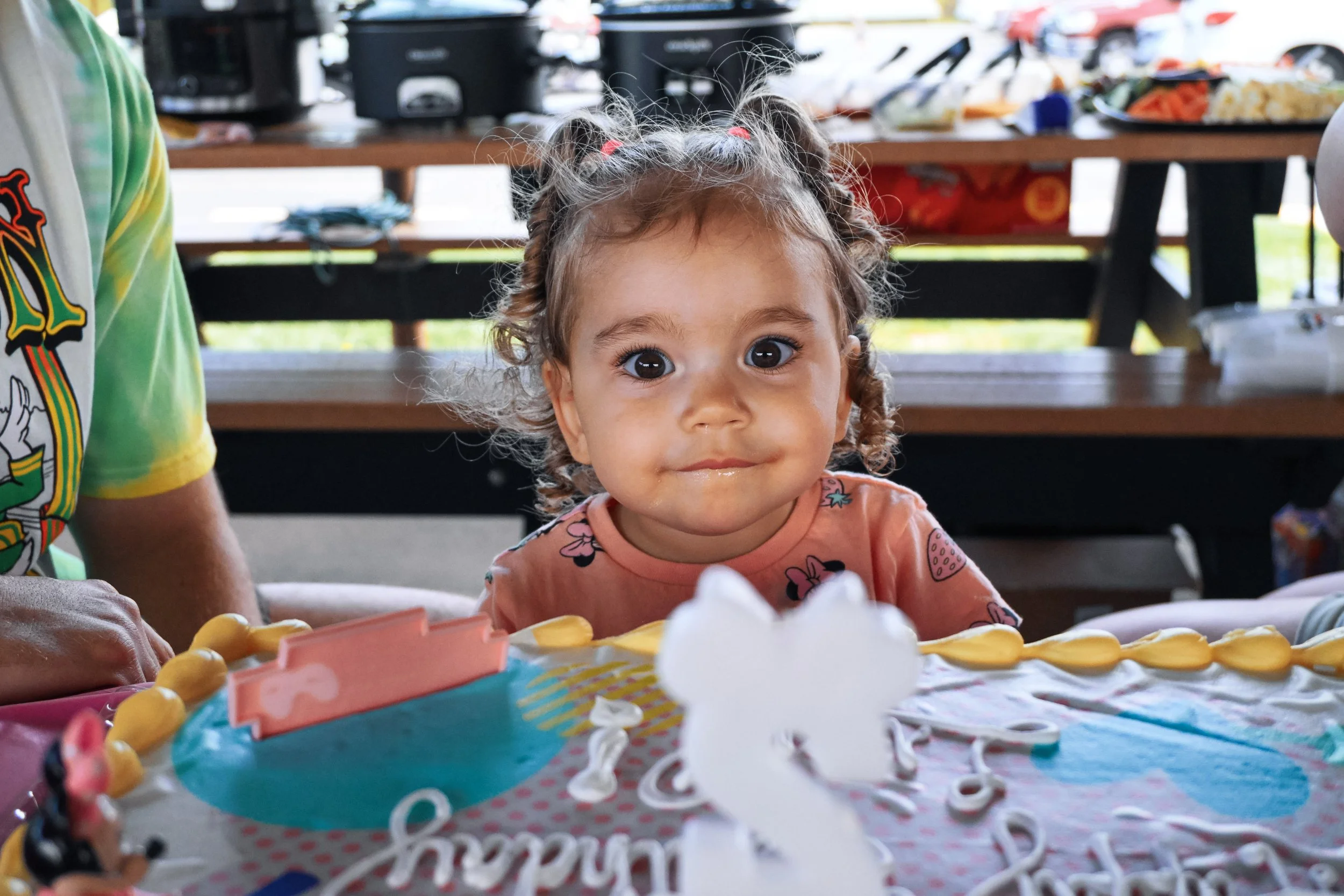 A young girl with curly hair tied in pigtails and wearing a pink shirt with cartoon characters, celebrates her birthday at a party with a decorated cake and colorful table setting. She looks directly at the camera with wide eyes and a slight smile in a festive outdoor setting.