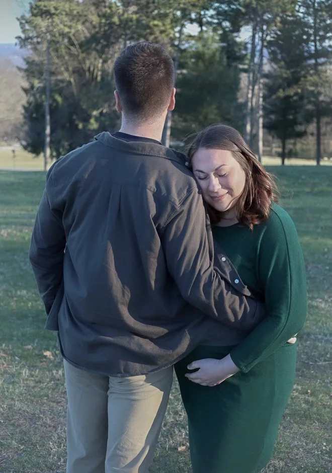 A couple standing outdoors in a park, embracing each other with the woman resting her head on the man's chest, both with closed eyes, surrounded by trees and grass.