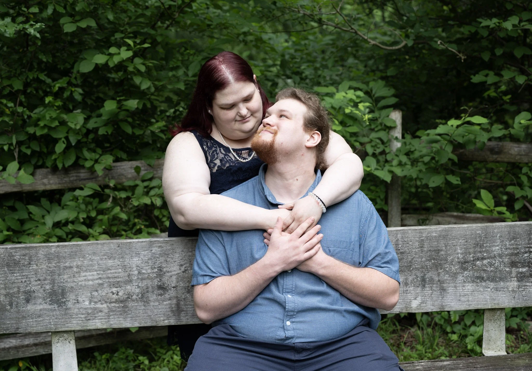 A woman with red hair embracing a man with glasses and a beard on a wooden bench in a green outdoor setting.