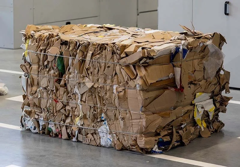 Large bundle of flattened cardboard boxes wrapped with string, placed on a floor in an indoor space.