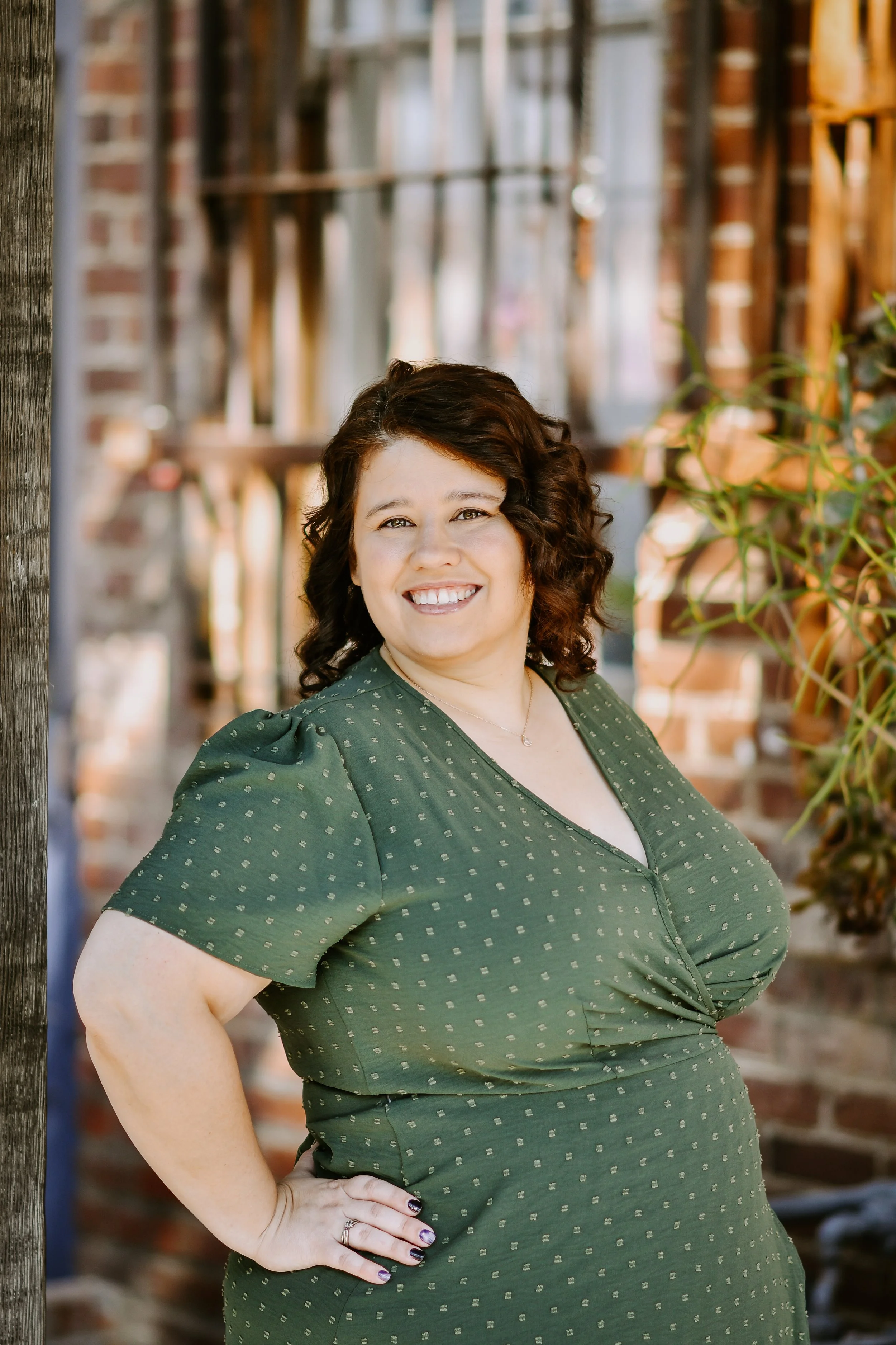 A woman with curly brown hair and a smile, wearing a green dress with small white polka dots, poses outside near wooden beams and brick walls.