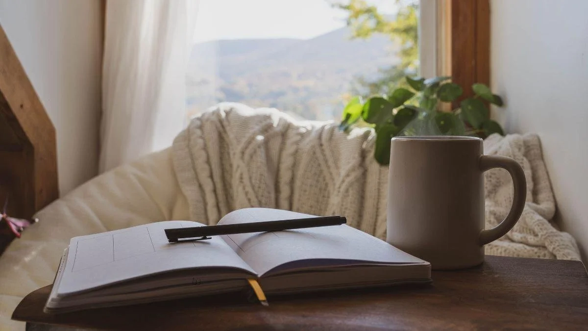 Open planner and black pen on a wooden table with a white mug, cozy blankets, and a plant by a window with a mountain view outside.
