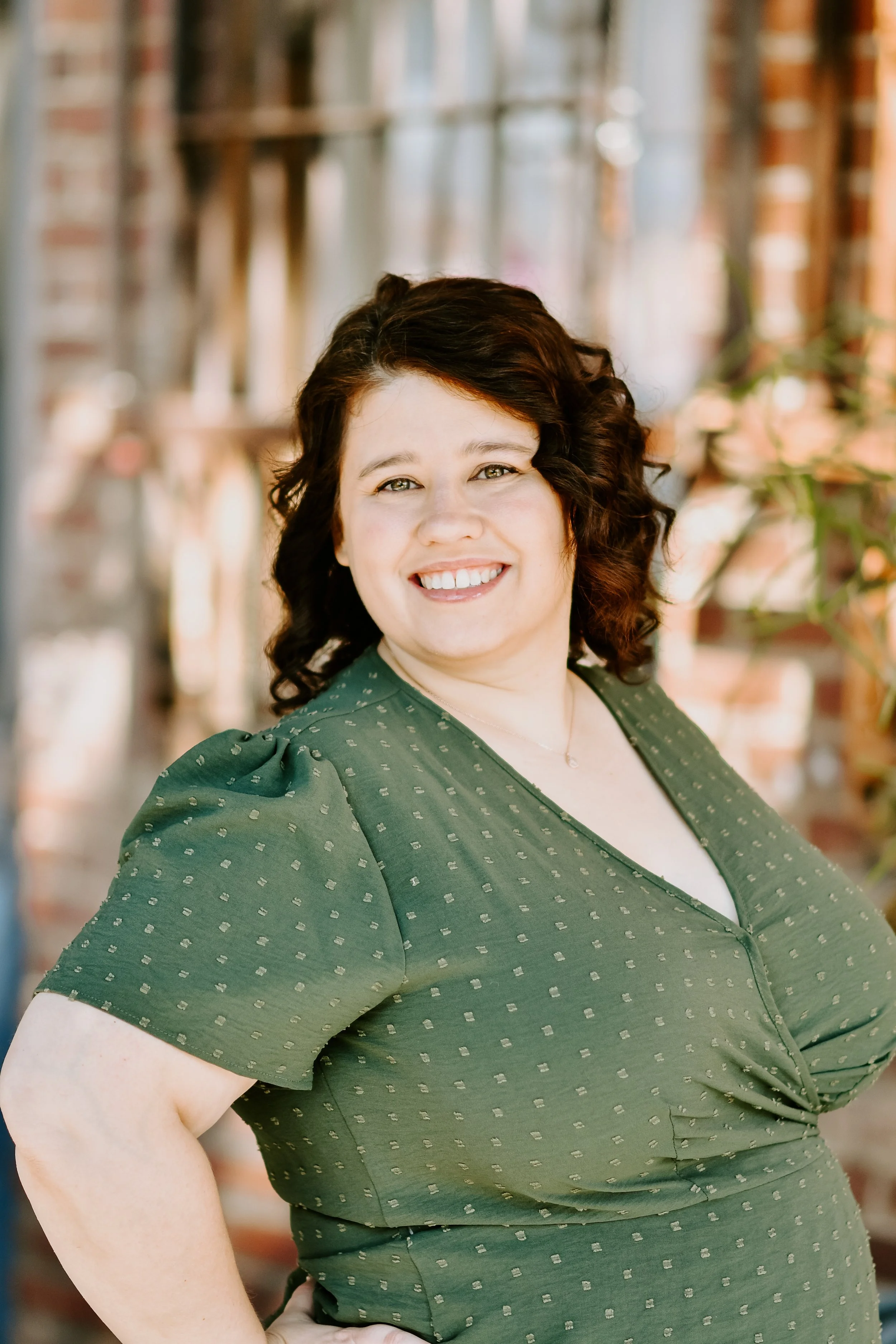 A woman with curly dark hair and light skin smiling, wearing a green dress with small dotted pattern, standing outdoors in front of a blurred background of a brick building with plants.
