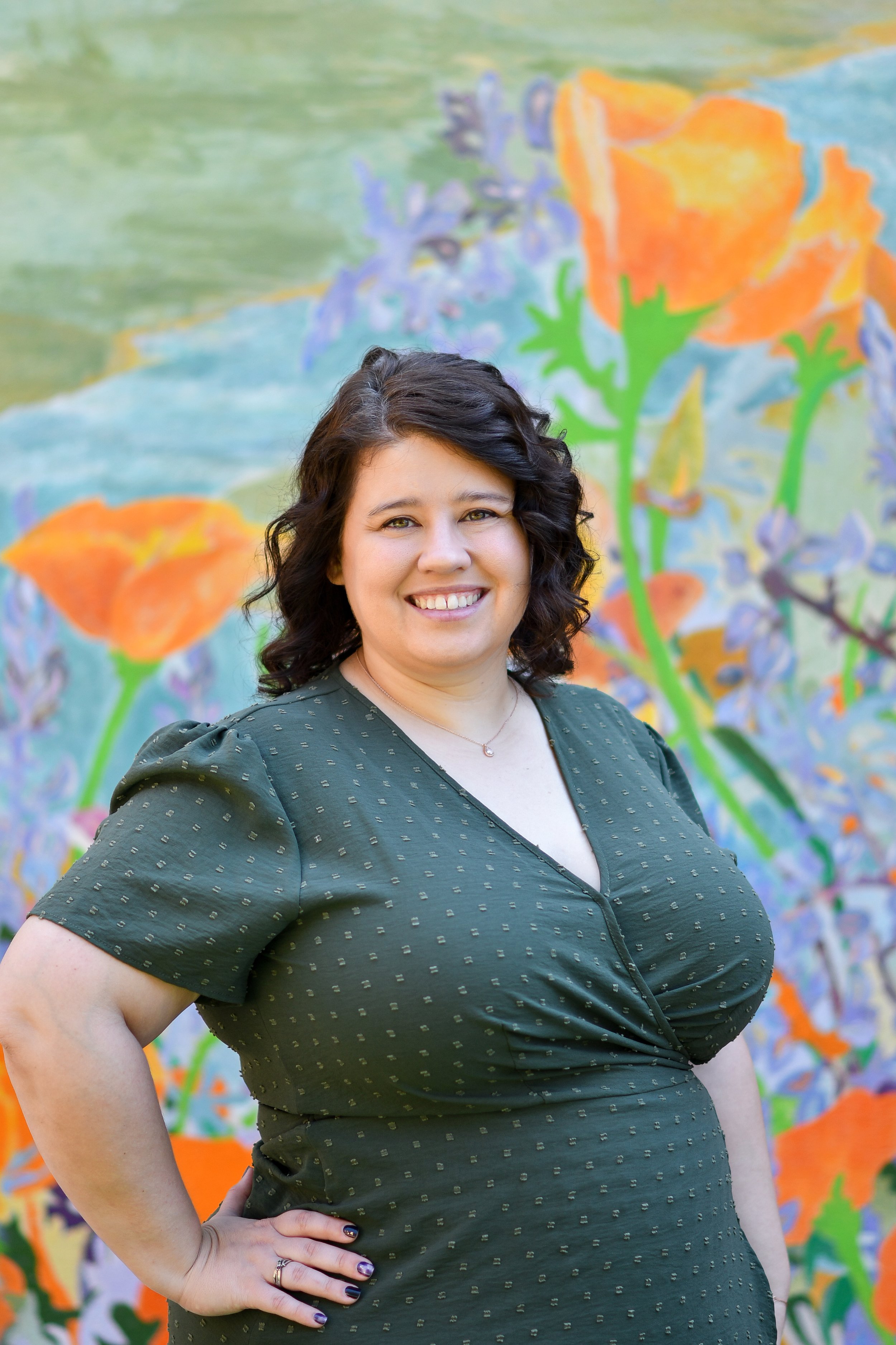 A woman with dark, curly hair smiling, standing in front of a colorful mural with orange flowers and purple accents.