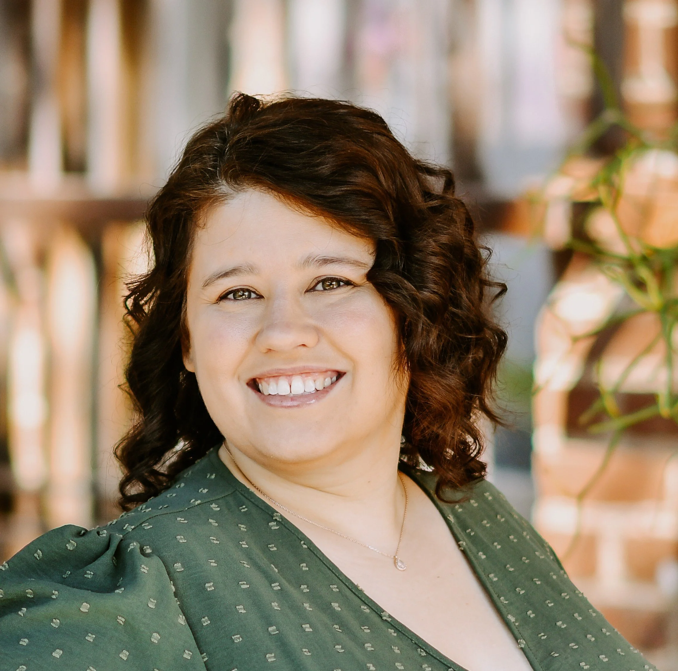 Close-up of a smiling woman's face with dark, curly hair, wearing a green dress with a dotted pattern, a necklace with a pendant, and a black strap across her shoulder, in front of a background with blurred vertical structures and plants.