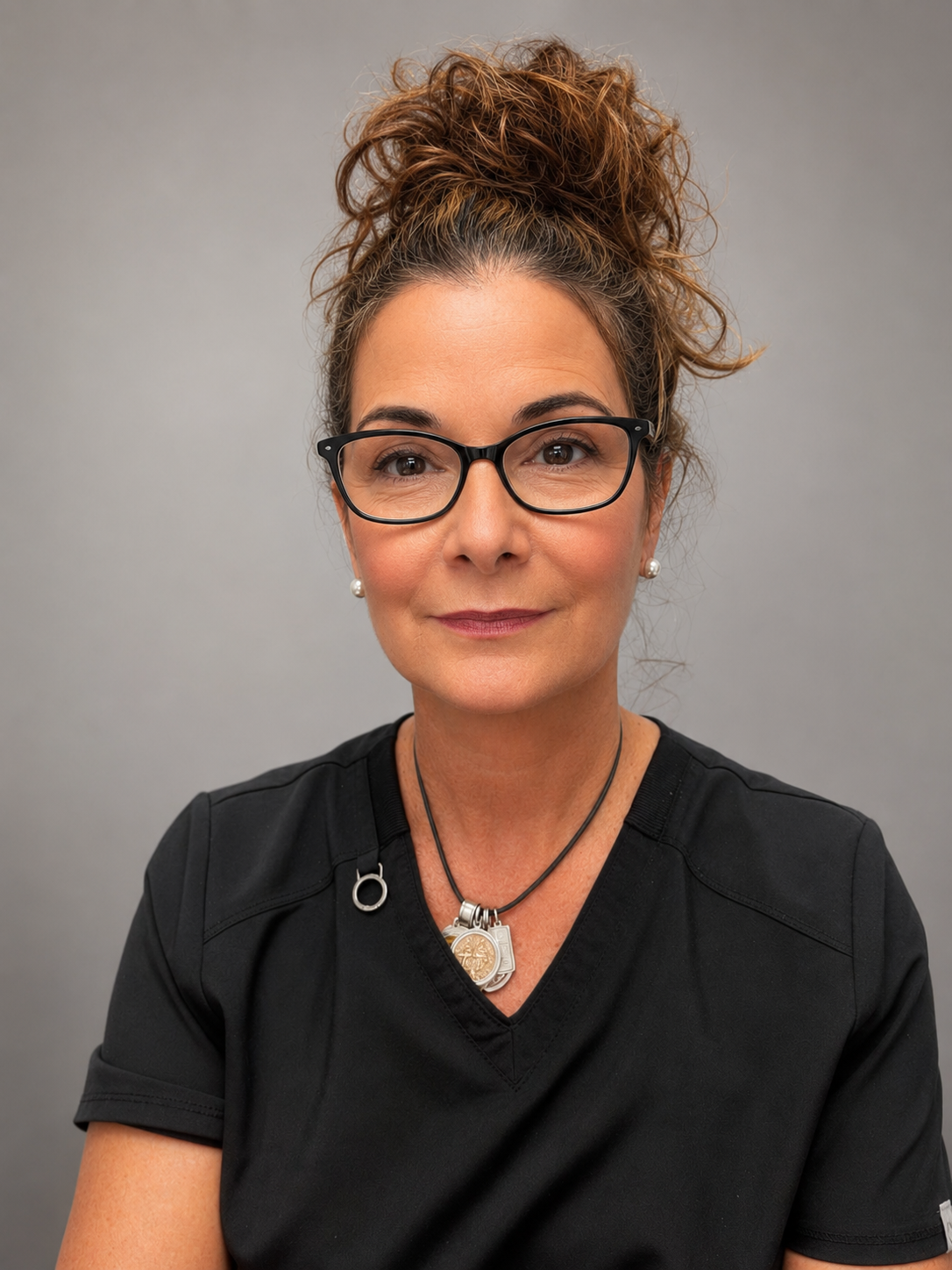 Portrait of Dr. Kate Crowley. She has curly brown hair in a high bun, is wearing black glasses, pearl earrings, and a black top, and standing against a gray background.