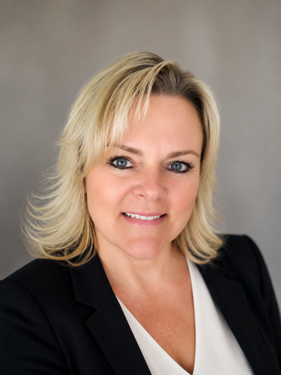 A portrait of a woman with blonde, shoulder-length hair, light blue eyes, wearing a black blazer and white top, smiling slightly against a neutral gray background.