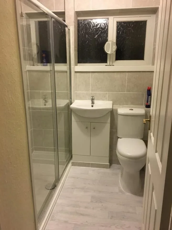 A small bathroom with a walk-in shower on the left, a white sink with a cabinet below in the center, and a toilet on the right. A window with frosted glass is above the sink and toilet, and a small bottle of cleaning spray is on the toilet tank.
