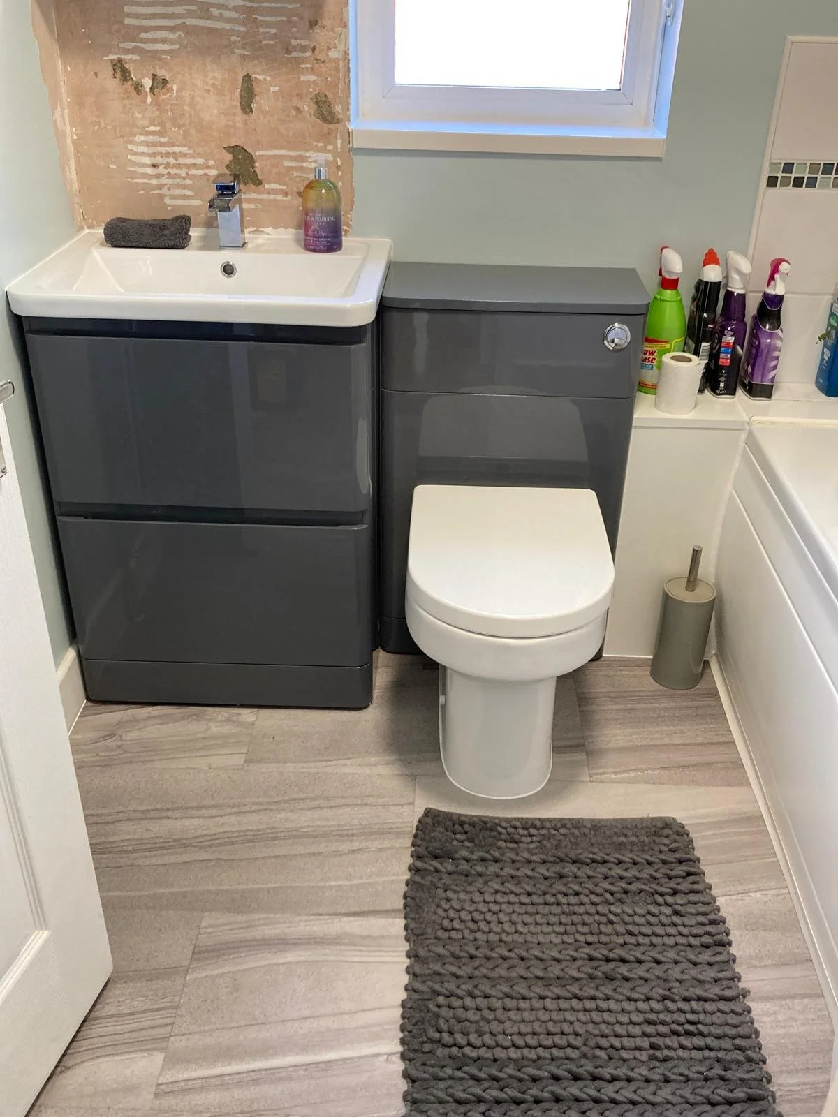 A compact bathroom with a washer and dryer, toilet, and laundry supplies. The washer and dryer are dark gray, with the washer on the left and the dryer on the right. The toilet is white, located between the appliances. There is a small gray rug in front of the toilet, and a paper towel roll on the floor near the toilet. On top of the dryer, there are personal care items, including spray bottles and a roll of toilet paper. The wall behind the washer and dryer has a window with frosted glass, and part of a bathroom wall with incomplete wallpaper or paint is visible.