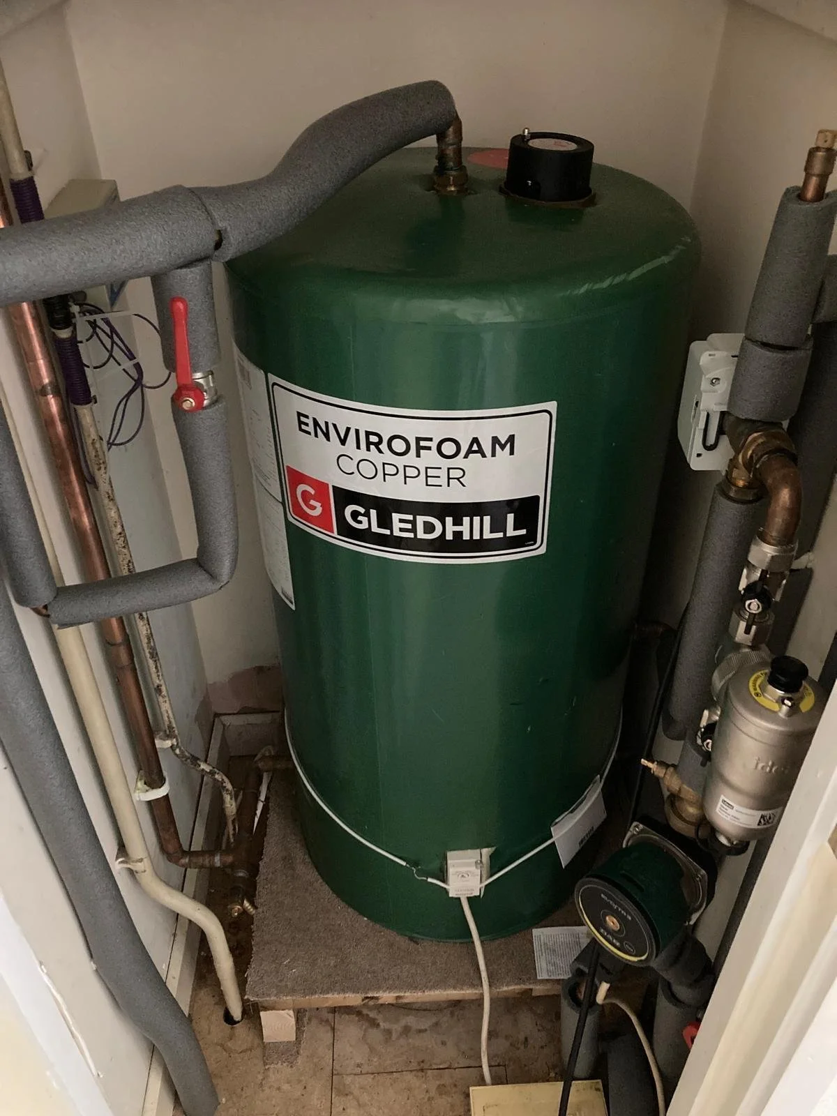 A green Gledhill Envirofoam copper hot water heater fitted in a closet with various pipes and a pressure gauge.