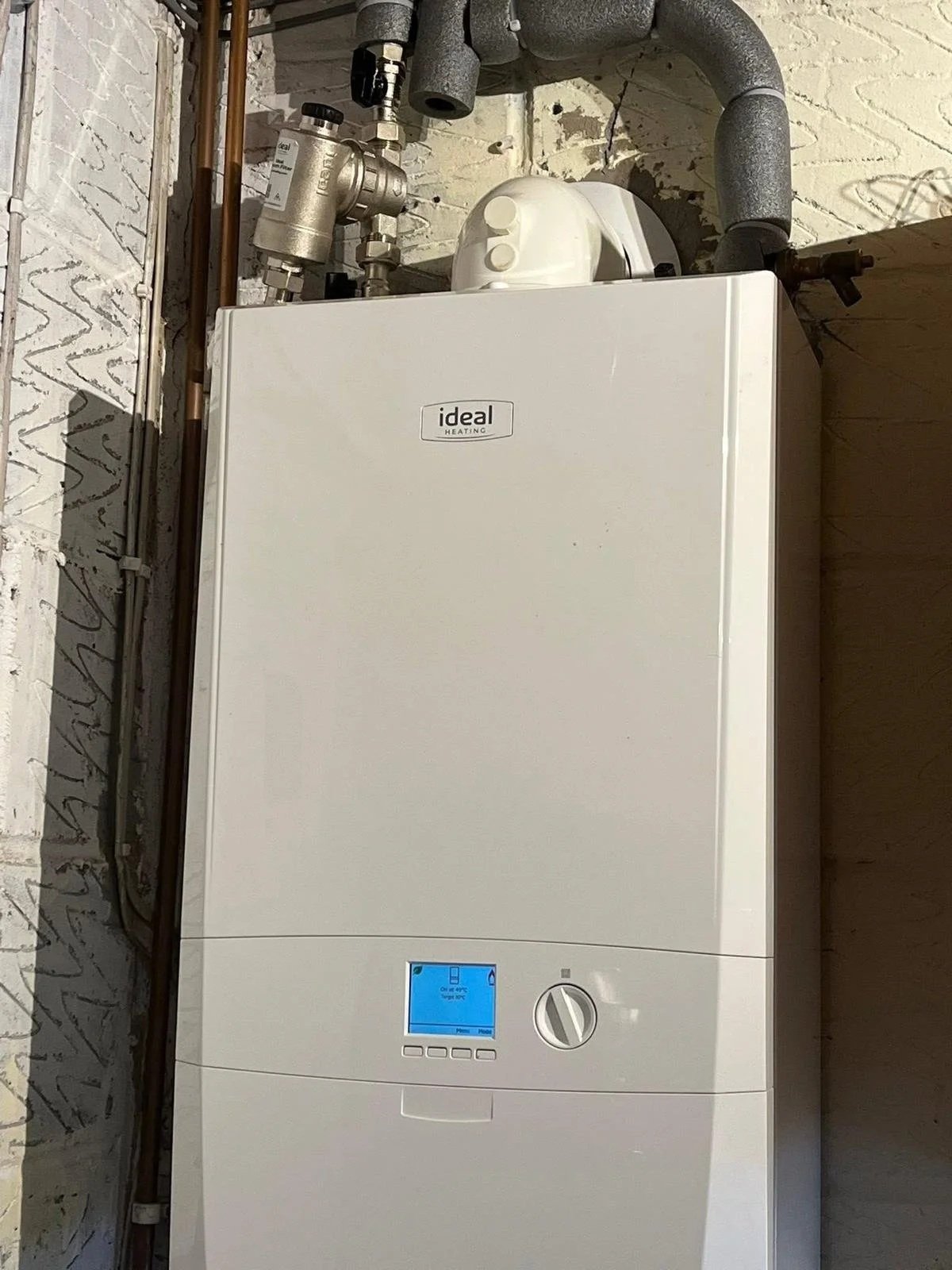 A white Ideal Heating boiler installed against a brick wall with visible piping and a digital control panel.