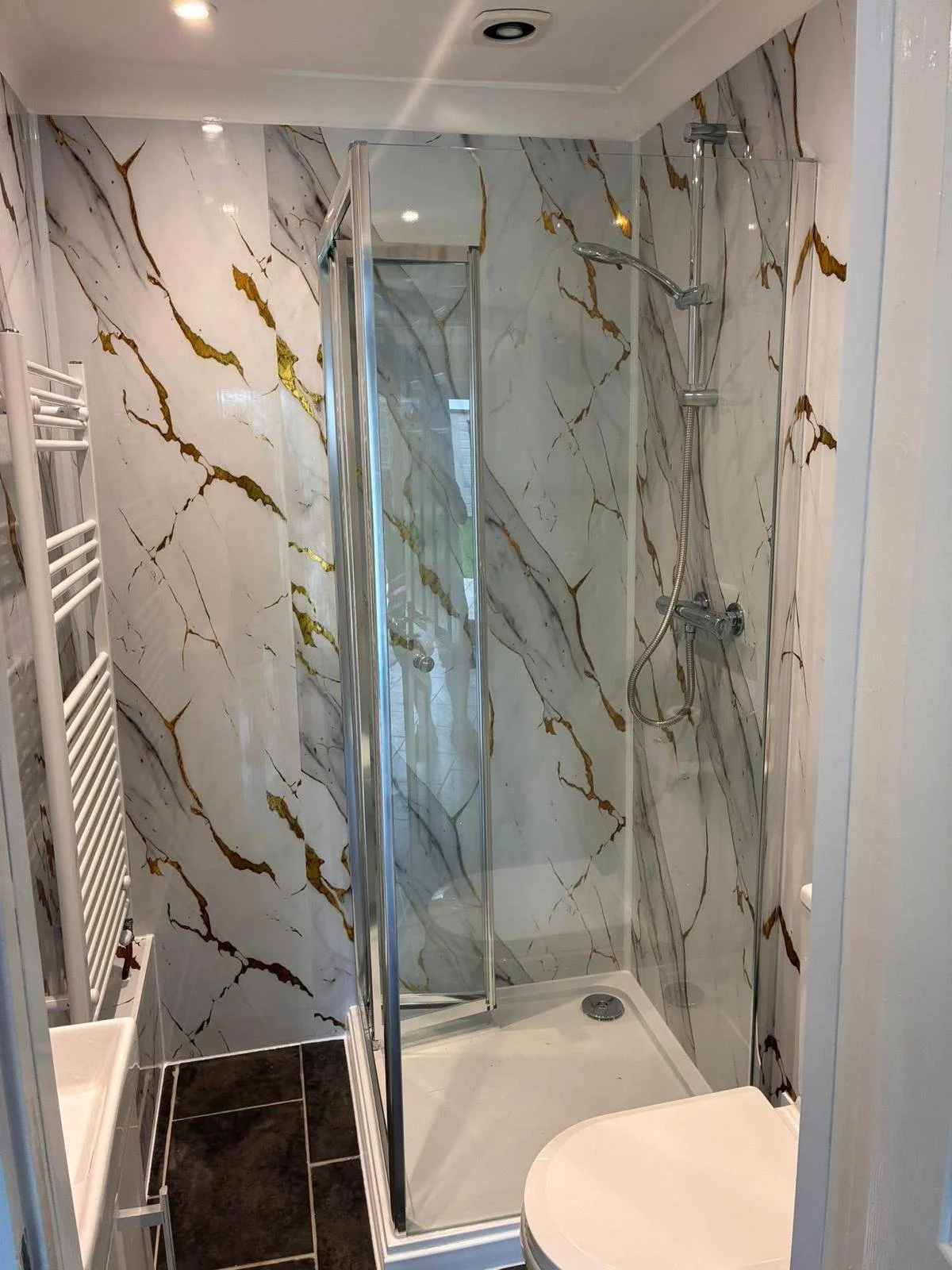 Small modern bathroom with marble-patterned walls, a compact shower stall with a glass door, a handheld showerhead, a white toilet, and a white towel radiator on the left.