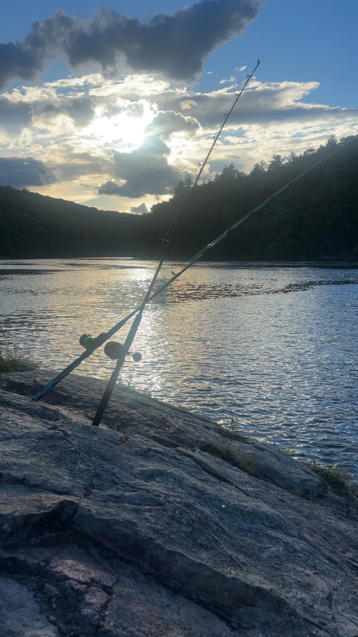 Two fishing poles resting on a rocky riverbank with a calm river, forested hills, and a cloudy sky with the sun partly obscured.