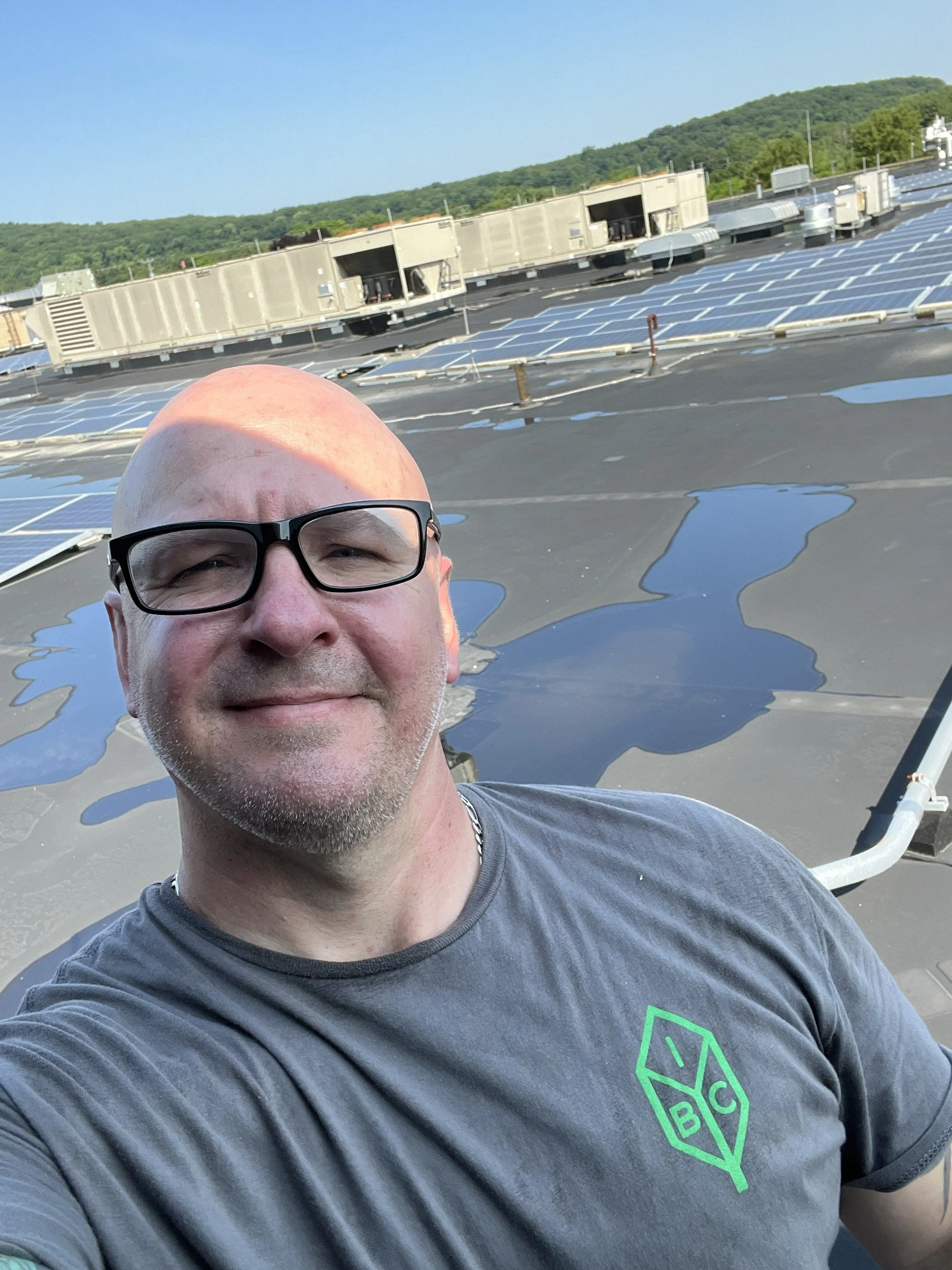 A man with glasses taking a selfie on top of a building with solar panels and HVAC units in the background, surrounded by a green landscape.