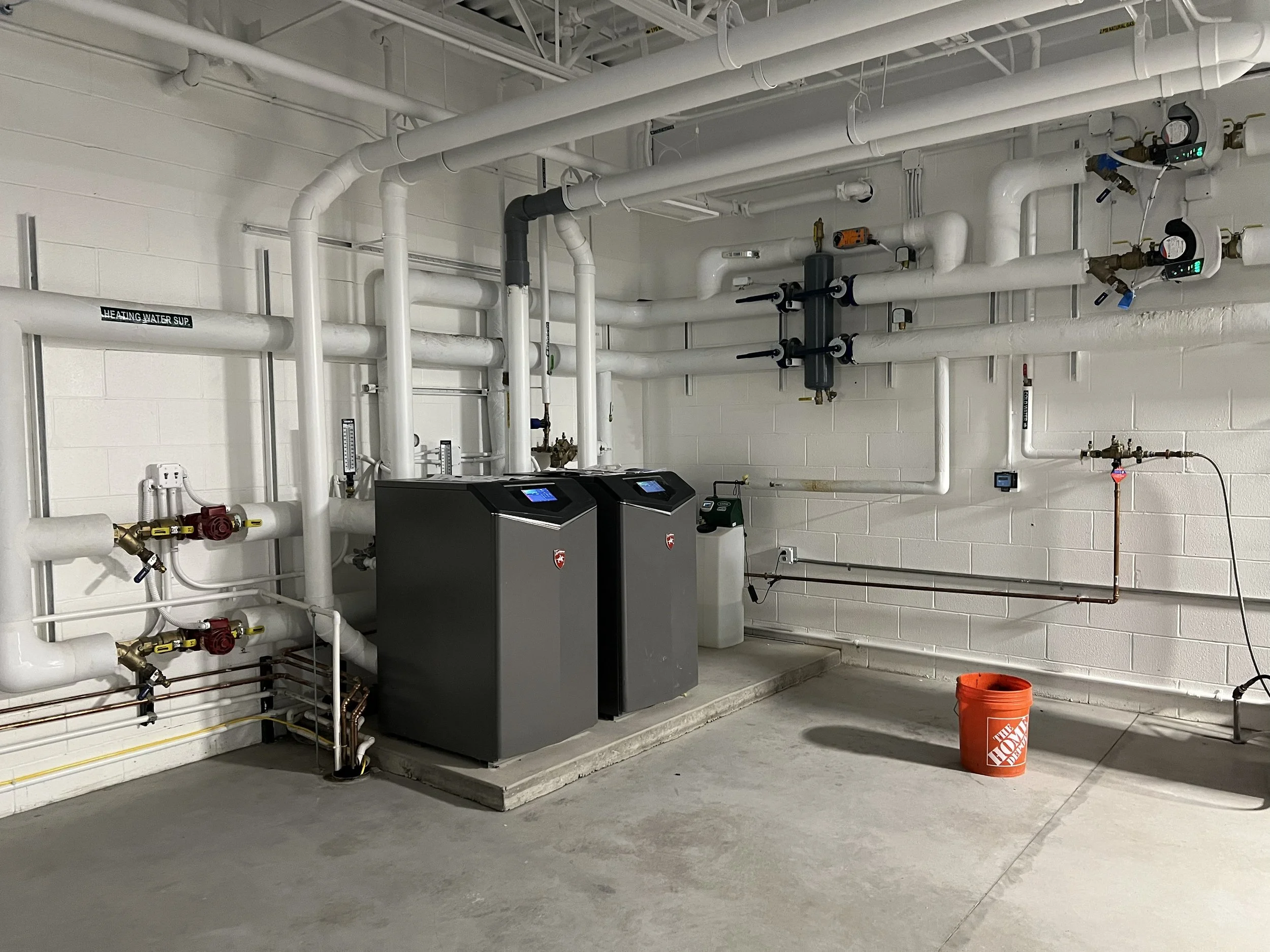 A mechanical room with white pipes, gauges, and equipment, including a home water heater and a red bucket on the floor.