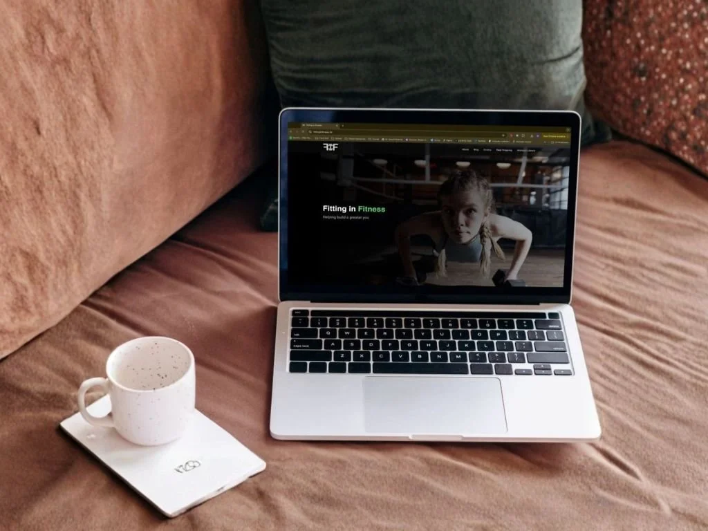 An open laptop on a brown couch with a white coffee mug and a notebook, displaying a website with a woman doing a workout.