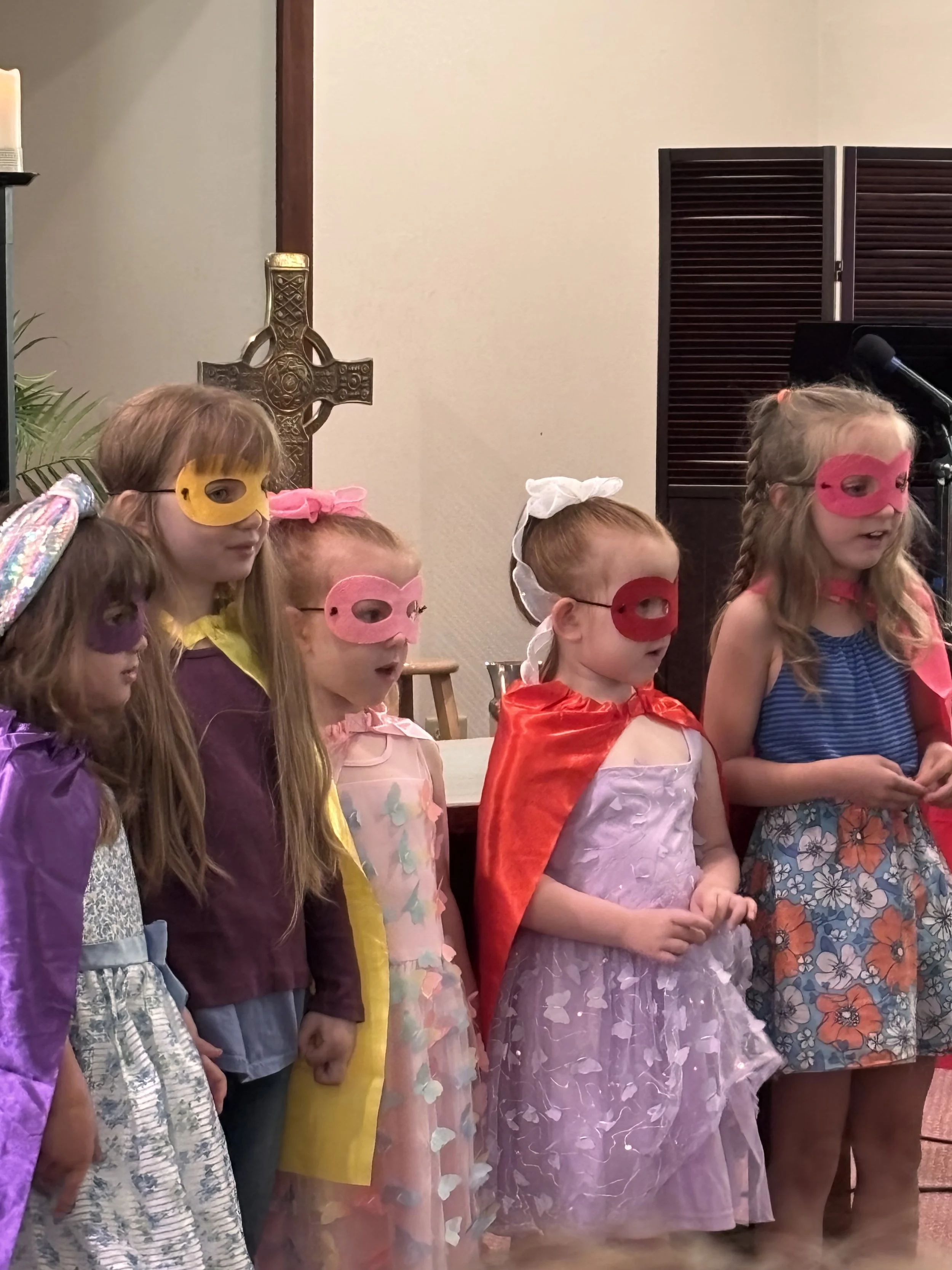 Children dressed in colorful costumes and masks, standing in a line during a performance or celebration in a church or community hall, with a cross and a speaker visible in the background.