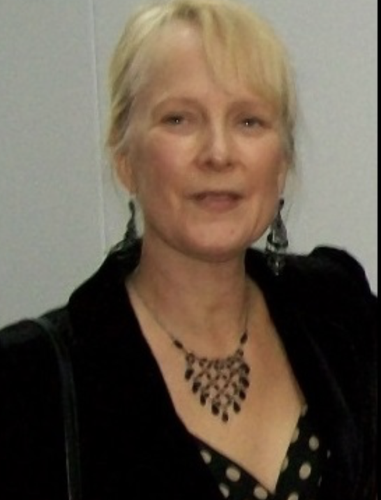 A woman with blonde hair smiling, wearing a black top with a polka dot pattern, a black jacket, and a necklace, standing against a plain background.