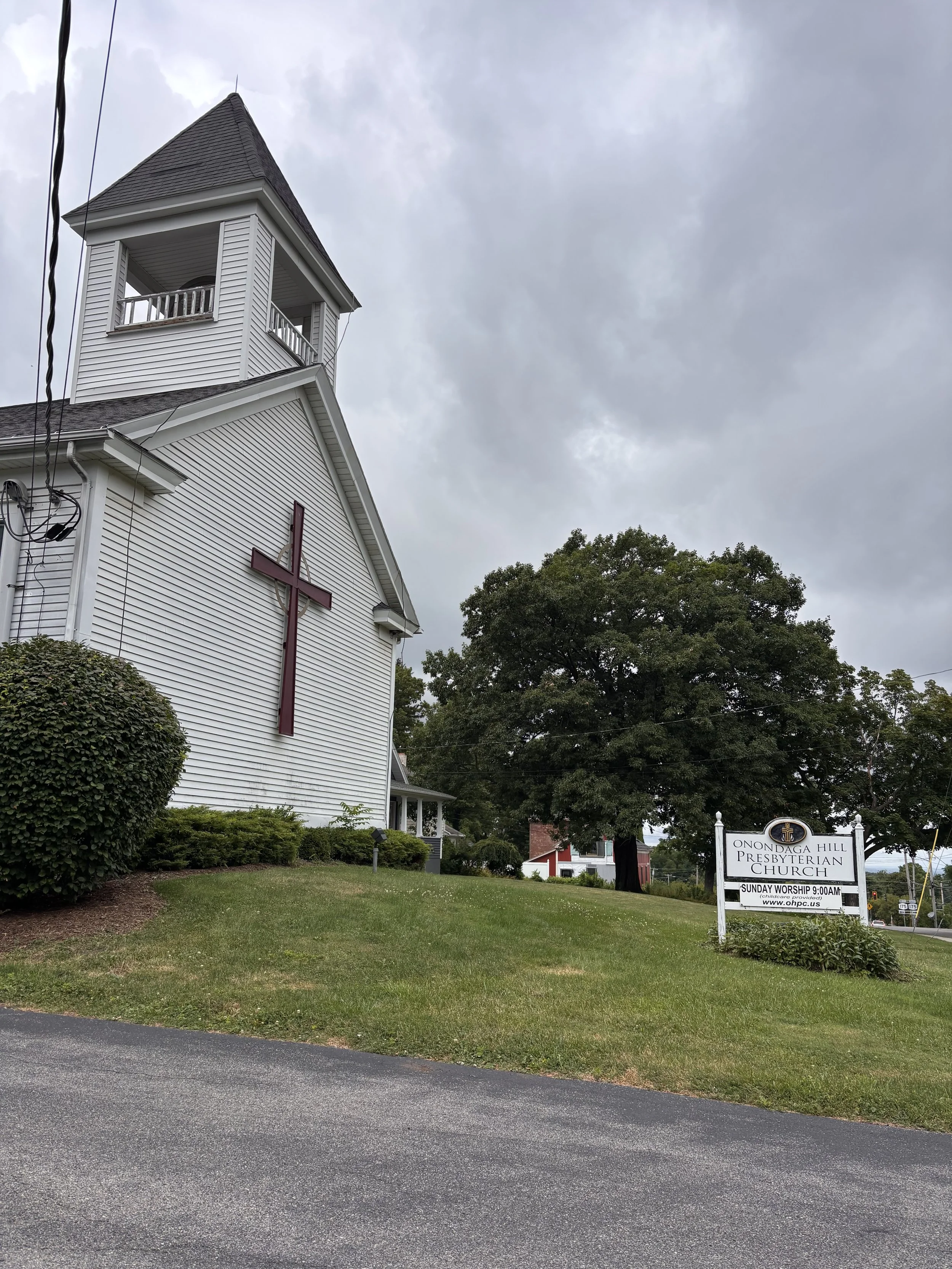 A white church with a red cross on the front, a tall steeple, and a sign outside reading "Onondaga Hill Presbyterian Church, Sunday Worship 9:00AM". The church is surrounded by green bushes and trees, with a cloudy sky overhead.