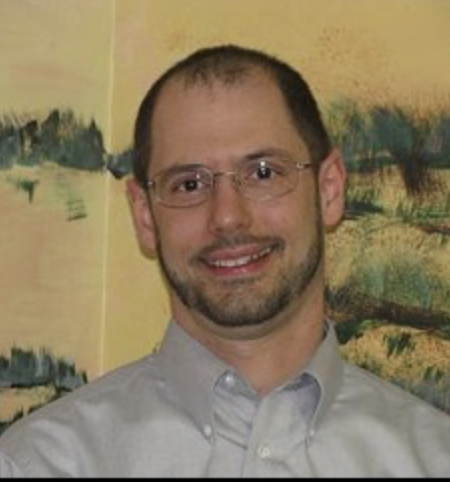 A man with short dark hair, glasses, and a beard, smiling, wearing a light gray button-up shirt, standing in front of a colorful abstract painting.