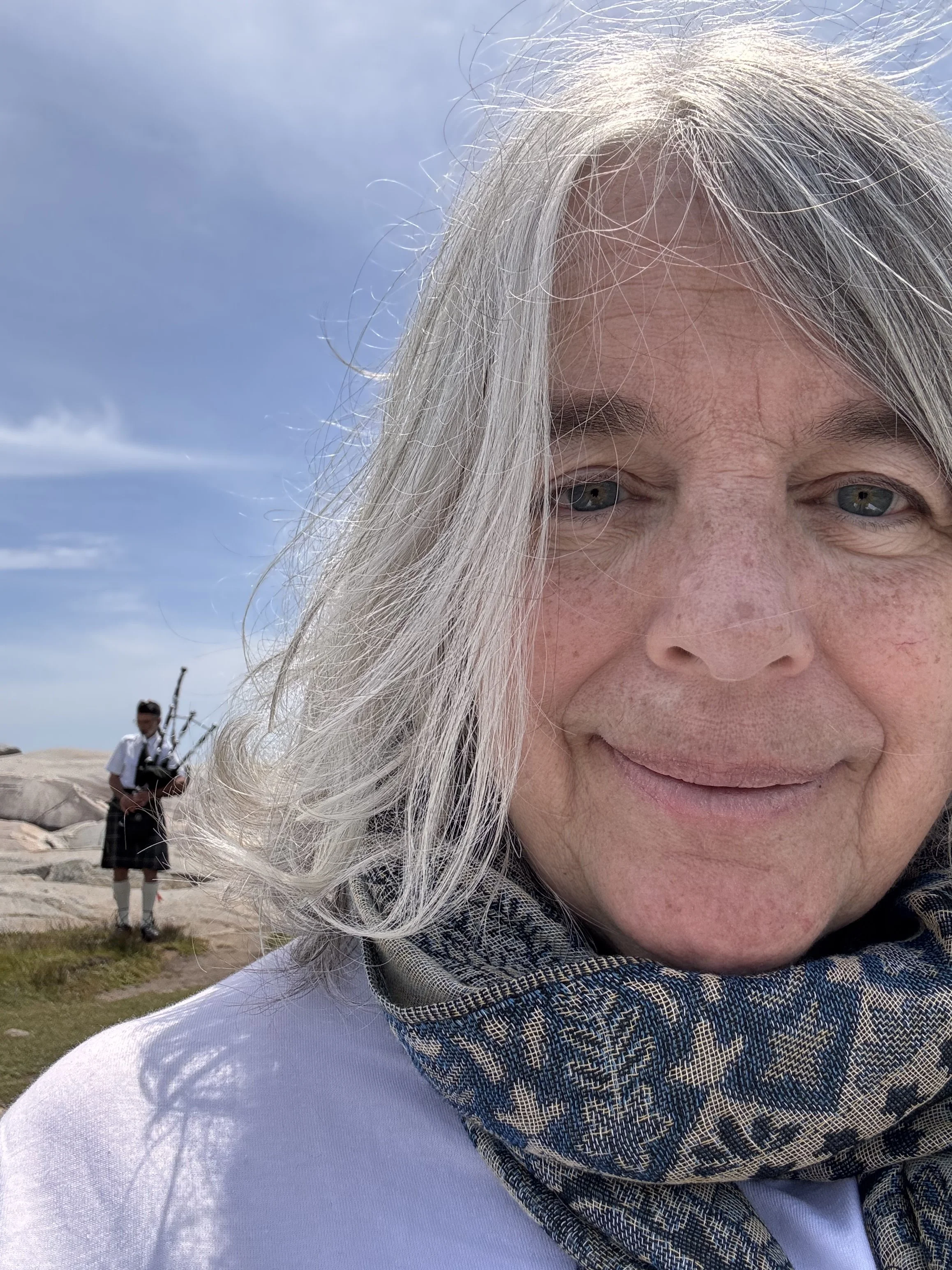 Close-up of an older woman's face with gray hair, blue eyes, and wearing a scarf, outdoors on a sunny day with a man fishing in the background.