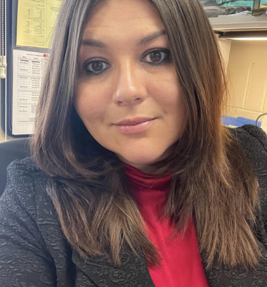 A woman with long brunette hair, wearing a black patterned jacket and a red top, sitting indoors with a neutral expression.