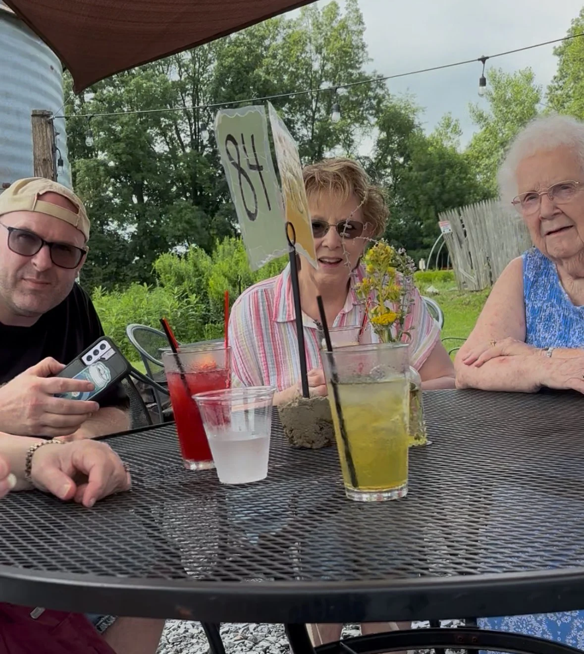 Four people sitting at an outdoor table with drinks, two women and one man holding a phone, surrounded by greenery with trees and a wooden fence in the background.