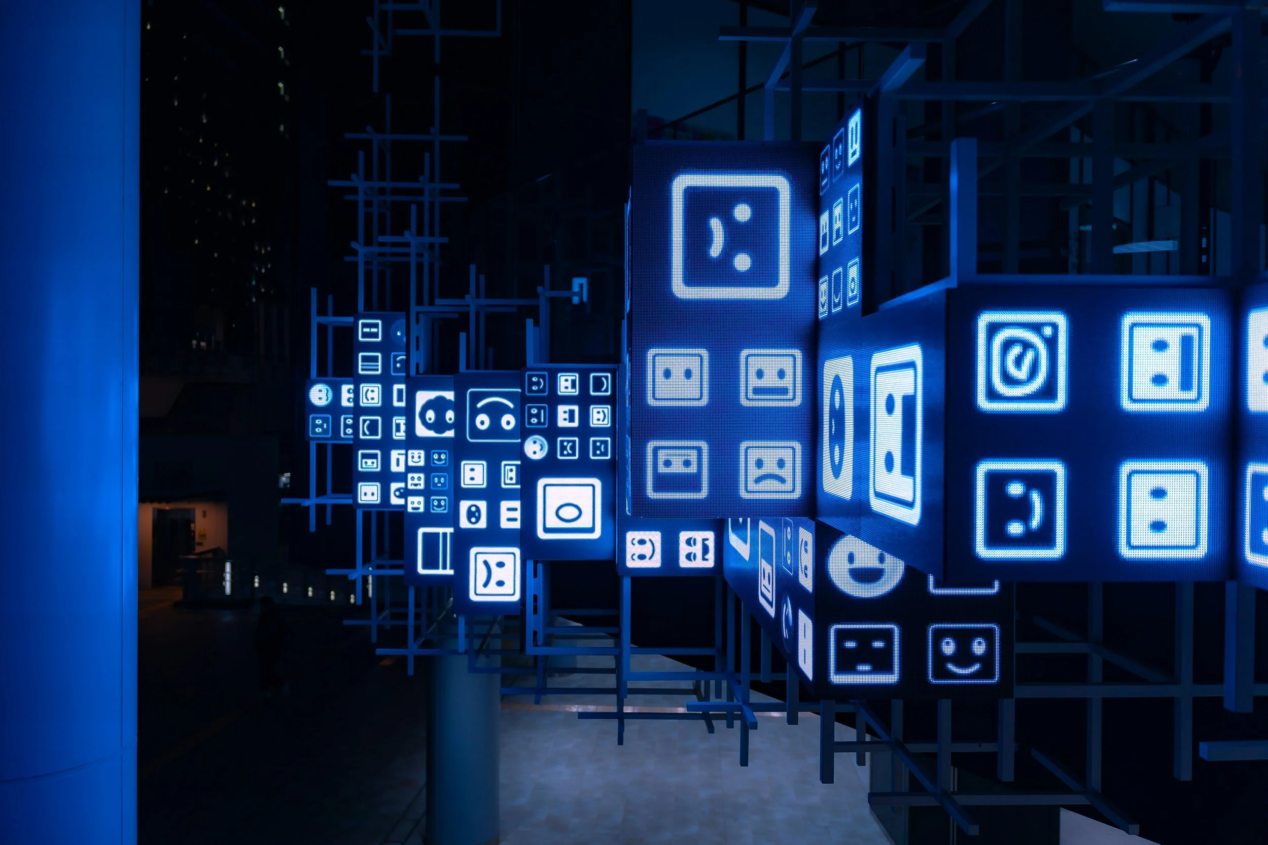 Nighttime city scene with an art installation featuring blue illuminated digital emojis and icons displayed on cubical structures arranged in a grid pattern.
