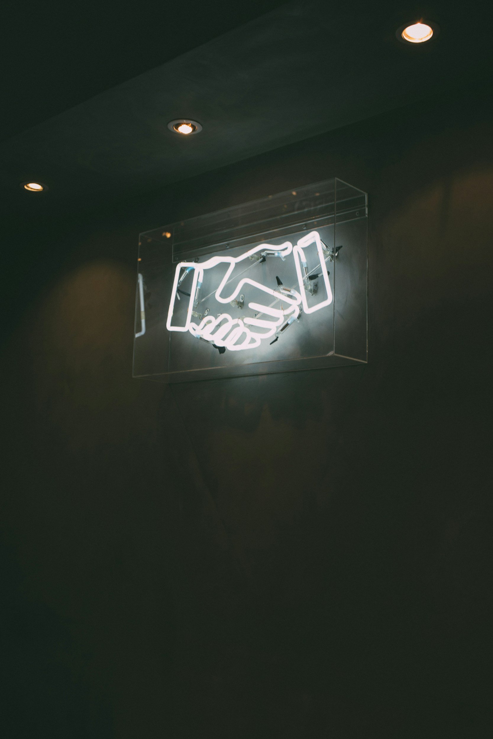 Neon sign depicting a handshake mounted on a dark wall, with a glass case around it and a ceiling with recessed lighting.