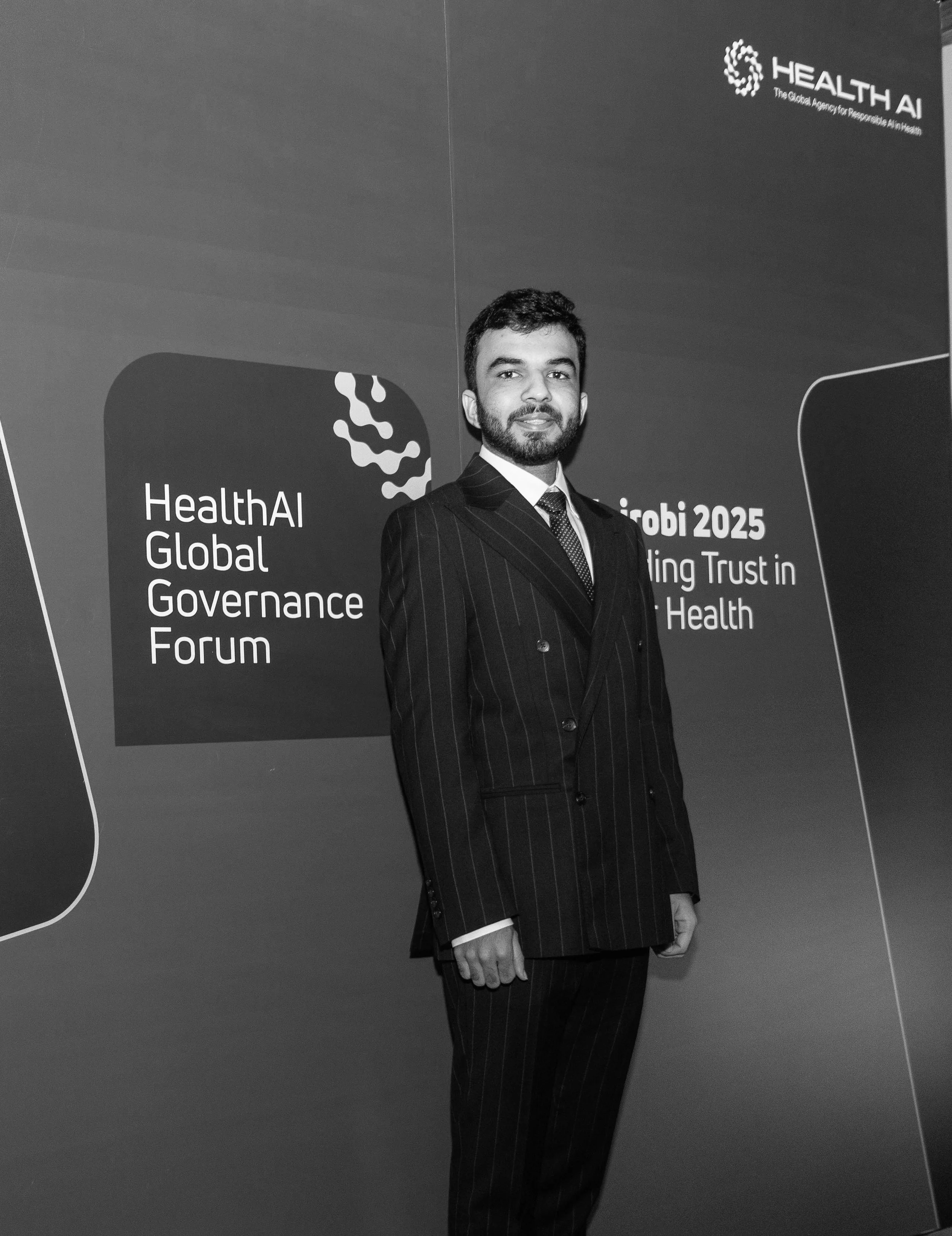 A man in a pinstripe suit standing in front of a large sign that reads 'Health AI Global Governance Forum' and 'Hirobi 2025: Building Trust in AI for Health'. The background is dark, and the person has short dark hair and a beard.