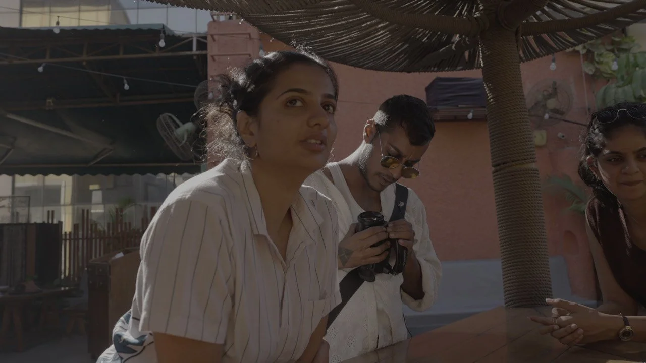 Three people standing outside under a thatched umbrella, one woman with curly hair in a striped shirt, a man with sunglasses looking at his phone, and another woman with sunglasses on her head and clasped hands.