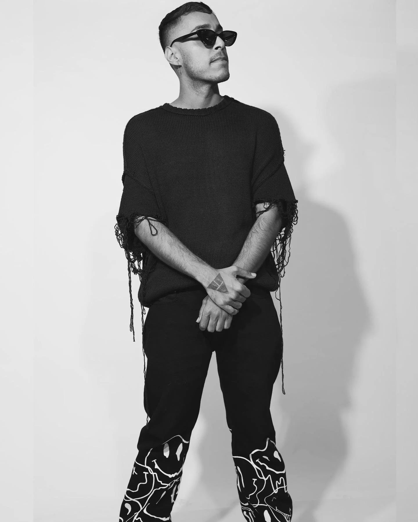 A man in black clothes, wearing sunglasses, with short hair, standing against a plain light background, looking sideways with arms crossed.