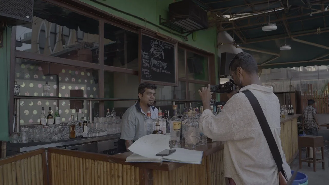 A man filming another man at a bar with bottles of alcohol behind the counter