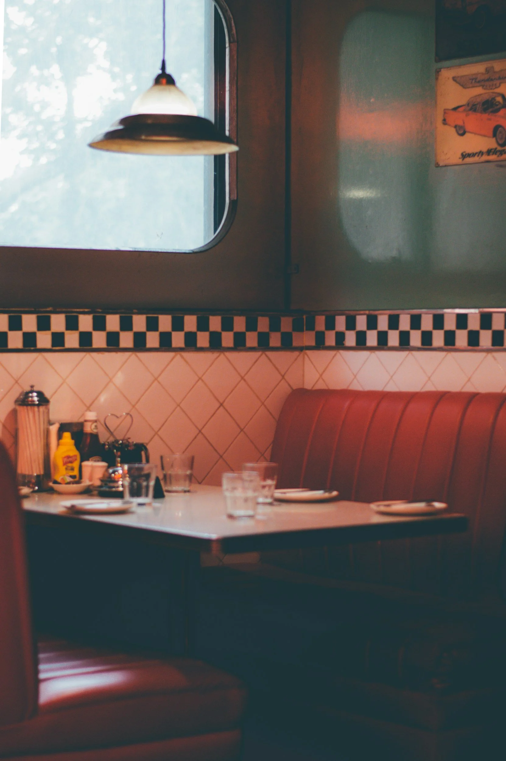A cozy retro-style diner with a table set for four, featuring glasses of water, plates, and condiments. A booth with red upholstery lines the wall. A window with a hanging lamp above it provides soft lighting. Decor includes vintage car signs and a checkerboard border.