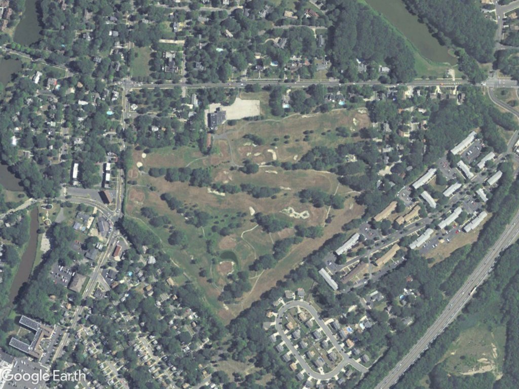 Aerial view of a park with open green spaces, trees, pathways, and surrounding residential areas with houses and apartment buildings.