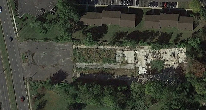Aerial view of an area with overgrown patch, driveway, and nearby parking lot, surrounded by trees and residential buildings.