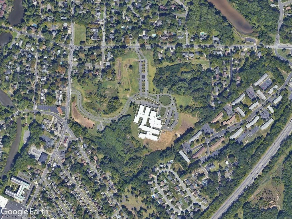 Aerial view of a suburban area with residential houses, roads, a large building complex in the center, and green spaces including trees and a pond.