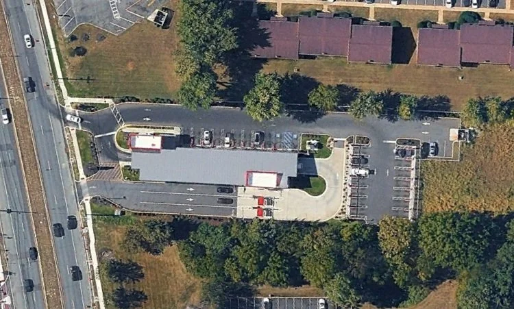 Aerial view of a parking lot and a building with a large gray roof, surrounded by trees and adjacent to a multi-lane highway.