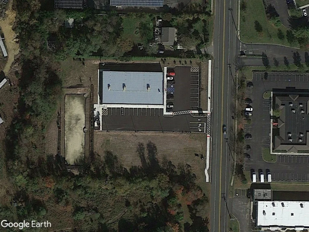 An aerial view showing a building with a parking lot, a road with cars, and surrounding greenery.