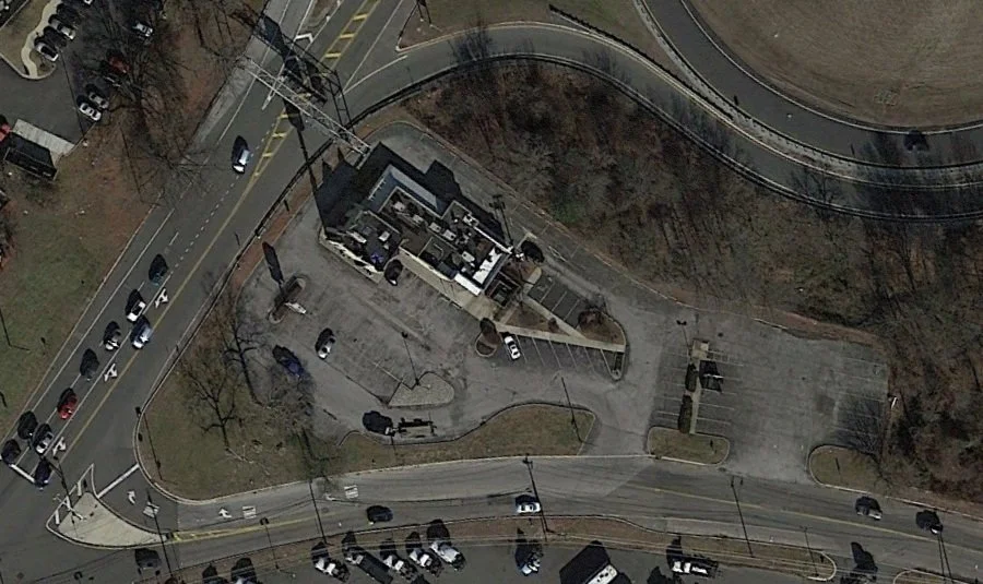 An aerial view of a parking lot and surrounding roads with parked cars and a small building.