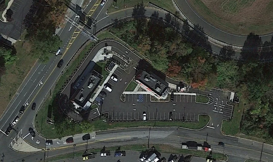 Aerial view of a parking lot adjacent to a multi-story building, surrounded by roads, trees, and power lines.